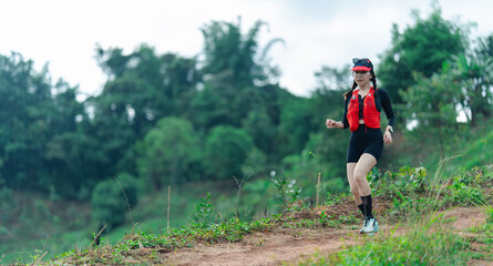 A woman wearing a red vest and black athletic outfit runs on a dirt trail surrounded by green trees...