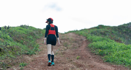 A person wearing athletic gear hikes up a dirt trail surrounded by green vegetation under a bright sky.