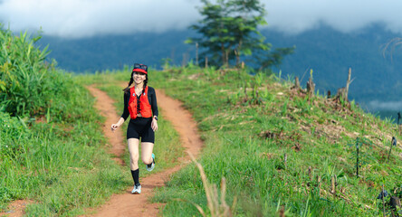 A woman jogs on a dirt trail surrounded by lush greenery with mountains and clouds in the...