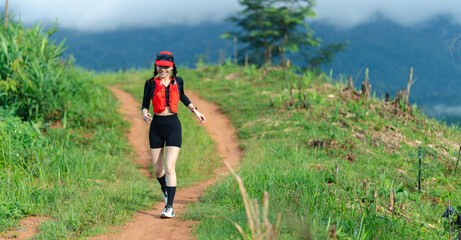 A runner wearing a red vest and headband jogs along a dirt path surrounded by green vegetation and...