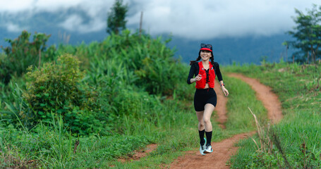 A woman in athletic gear runs on a dirt path surrounded by lush greenery and misty mountains in the...