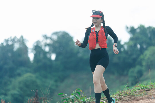 A woman wearing a red hydration vest and visor runs on a trail surrounded by greenery and trees.