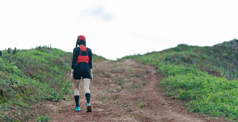 A person hiking uphill on a dirt path surrounded by green vegetation under a cloudy sky.
