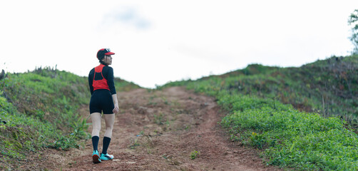 A person in athletic gear walks up a dirt trail surrounded by green vegetation under a cloudy sky.