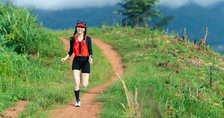 A woman jogs along a dirt trail surrounded by lush greenery and hills under a cloudy sky.