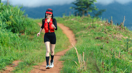 A woman in athletic gear runs on a dirt trail surrounded by green grass and hills under a cloudy sky.