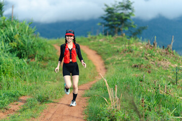 A woman in athletic gear runs along a dirt path surrounded by green grass and hills under a cloudy...