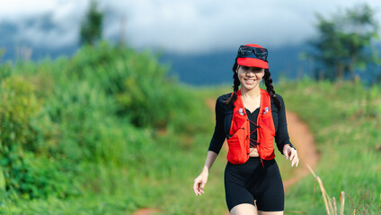A woman wearing a red life jacket and black outfit walks along a green trail with a scenic, cloudy mountain backdrop.