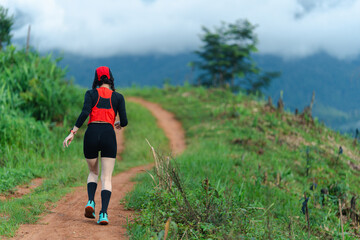 A person wearing athletic gear jogs on a dirt trail winding through green hills under a cloudy sky.