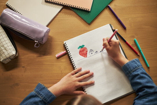 Child drawing strawberry and houses in notebook using colored pencils, hands visible on desk with school supplies, Caucasian girl creating artwork during creative activity