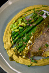 An elegant plate featuring delicate roasted fish served atop a bed of smooth yellow puree, garnished with vibrant green asparagus, edamame, and microgreens showcasing culinary art.