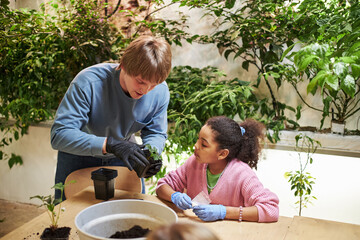 Caucasian young man demonstrating plant care to Black girl at table, both wearing gloves,...