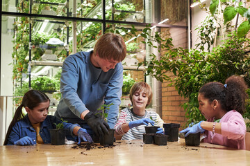 Caucasian young man teaching three children of diverse ethnicities gardening skills indoors, all...