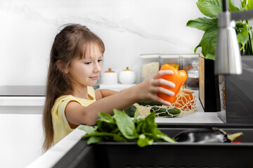A little girl takes out organic vegetables and fruits from a mesh bag in the modern kitchen. Grocery shopping. Zero Waste. Eco-Friendly Shopping. The concept of healthy lifestyle