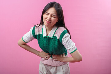 Asian woman clutching her stomach with a pained facial expression, suggesting abdominal discomfort or stomachache, standing in front of a bright pink background