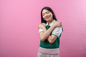 Asian woman closing eyes and smiling sweetly while hugging herself with both arms, wearing a green...