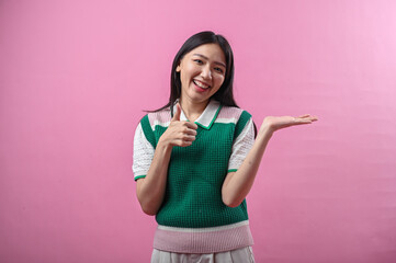 Asian woman smiling while pointing to her open palm, wearing a green knit vest and standing against...