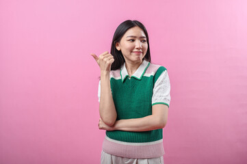 Asian woman in green knit vest pointing her thumb over her shoulder with a doubtful smirk, posing against a solid pink background