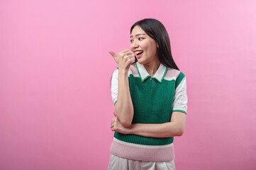 Asian woman in green knit vest pointing her thumb over her shoulder with a doubtful smirk, posing against a solid pink background