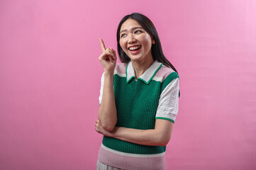 Asian woman looking up while smiling and pointing her index finger upward with an inspired expression, standing against a plain pink background