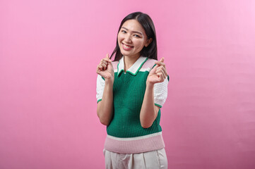 Asian woman smiling with both hands making Korean finger heart gesture, wearing a green knit vest, standing against a solid pink background