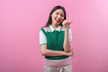 Asian woman in green knit vest pointing her thumb over her shoulder with a doubtful smirk, posing...