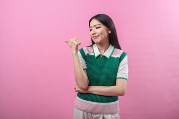 Asian woman in green knit vest pointing her thumb over her shoulder with a doubtful smirk, posing...