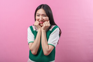 Asian woman with a playful expression poses with clenched fists under her chin, wearing a green...