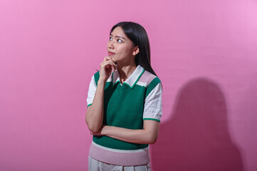 Asian woman in a green and pink sweater vest with a thoughtful expression, eyes closed and hand on chin, standing in front of a plain pink background