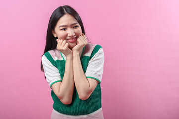 Asian woman with a playful expression poses with clenched fists under her chin, wearing a green...
