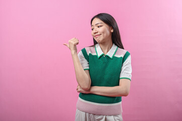 Asian woman in green knit vest pointing her thumb over her shoulder with a doubtful smirk, posing...