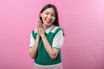 Asian woman clapping her hands and smiling warmly, wearing a green and pink knit top, standing...