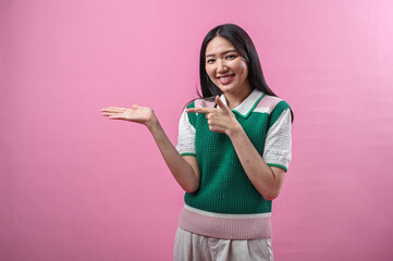 Asian woman smiling while pointing to her open palm, wearing a green knit vest and standing against...