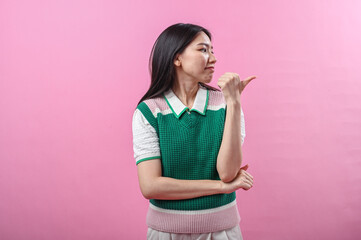 Asian woman in green knit vest pointing her thumb over her shoulder with a doubtful smirk, posing against a solid pink background