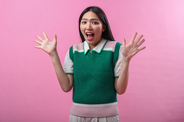 Asian woman in green sweater vest with joyful wide smile, raising both hands in excitement,...