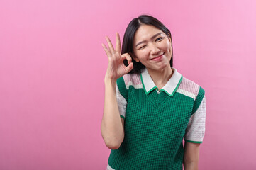 Asian woman winking and smiling while showing an OK hand gesture with right hand, posing...