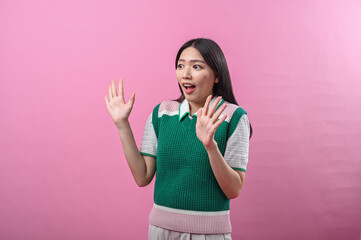 Fototapeta premium Asian woman in green knit top looking surprised and excited with both hands raised near her face, standing in front of a bright pink background