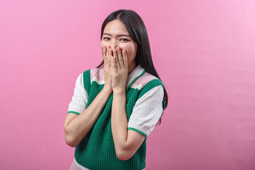 Asian woman in green vest expressing surprise with hands covering mouth, standing against a vibrant...