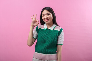 Asian woman wearing a green and pink knit vest smiling confidently while making an OK hand gesture...