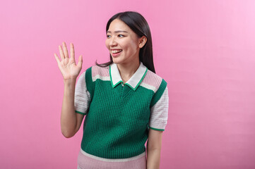 Asian woman waving and smiling in a friendly gesture, wearing a green and pink sweater with short...