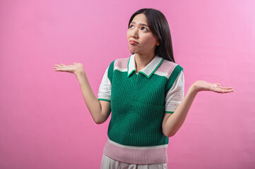Asian woman shrugging with confused expression, wearing green and pink sweater vest, arms raised,...