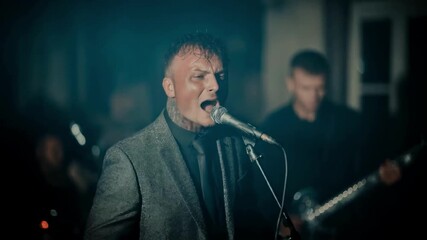 Passionate male rock singer in a suit performing emotionally into a microphone with his band on a dark stage, representing live music, concerts and the entertainment industry