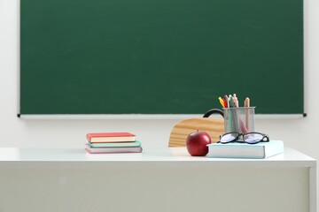 Different school stationery, glasses and red apple on white table near blackboard indoors