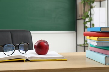 Different books, glasses and red apple on wooden table near blackboard indoors