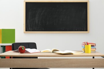 Different stationery and red apple on wooden table near blackboard indoors
