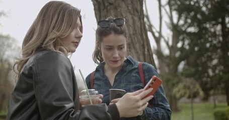 Two women enjoy coffee and conversation in the park. One woman shows her friend something interesting on her phone. - Powered by Adobe