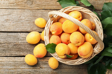 Fresh apricots in wicker basket and green leaves on wooden table, flat lay. Space for text