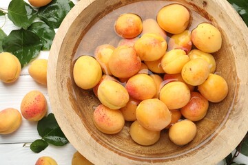 Fresh ripe apricots in bowl with water and green leaves on white wooden table, flat lay