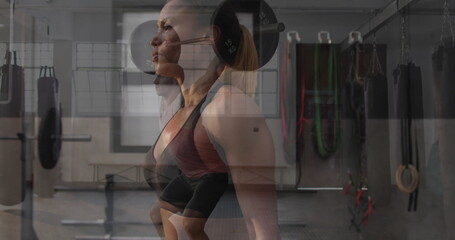 Lifting woman in tank top, leggings pressing barbell overhead in gym, with punching bags and rings
