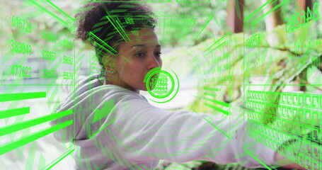 Woman wearing hoodie using holographic grids in garden, extending arm toward numeric code panels